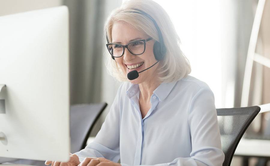 A mature woman working as a patient transfer support specialist discusses a case through her headset while typing notes.