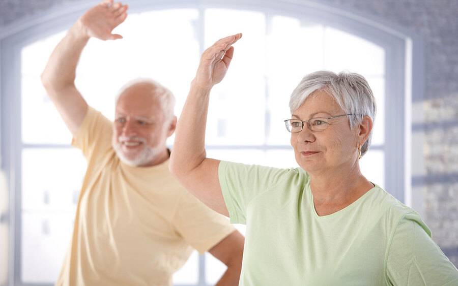 A senior man and woman stretch their arm up in an exercise class designed to improve circulation and mobility.