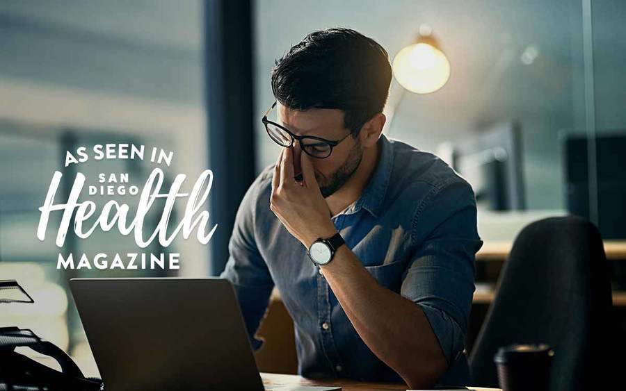 A young man sitting at a desk with his fingers pinching his nose represents how too much time sitting can hurt your health.