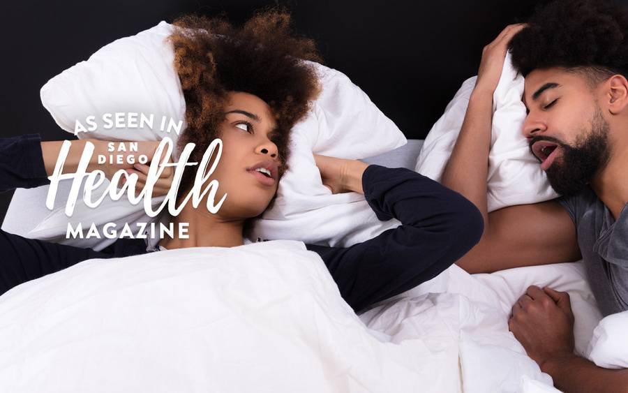 A young woman covers her ears with her pillow as she looks at her significant other snore as he sleeps. San Diego Health Magazine