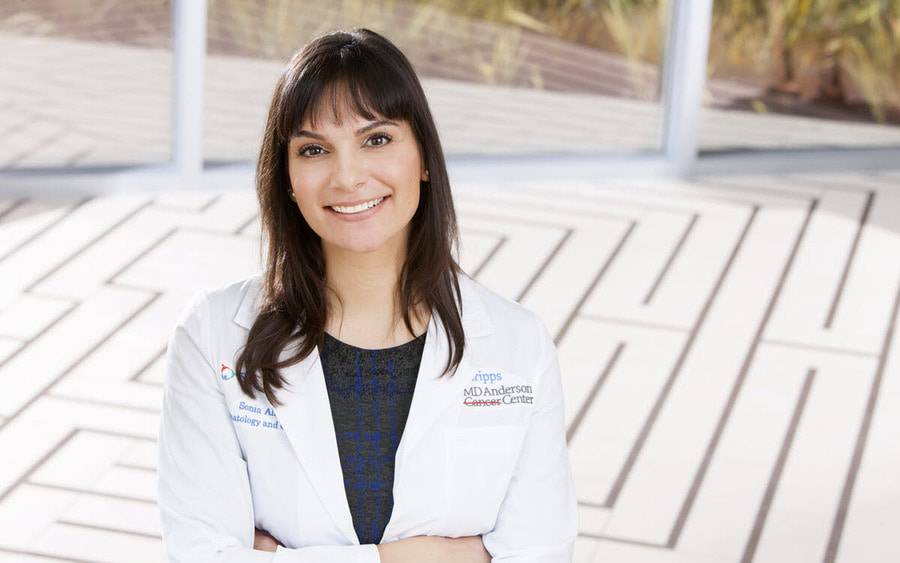 Sonia Ali, a breast medical oncologist with Scripps MD Anderson Cancer Center and is a Scripps Clinic physician, is smiling outdoors with her white coat. 