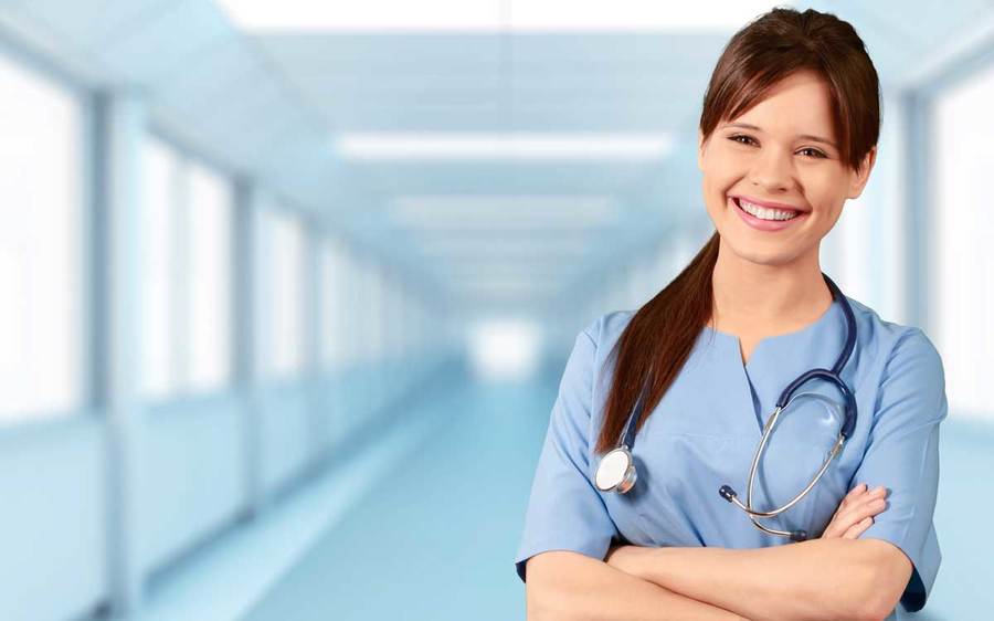 A smiling health care professional wearing scrubs and a stethoscope pauses in the hallway of a hospital