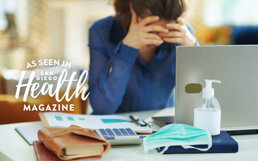 A woman stressed sitting at her at home desk with a mask and hand sanitizer near by, illustrating how coronavirus anxiety is effecting this woman.