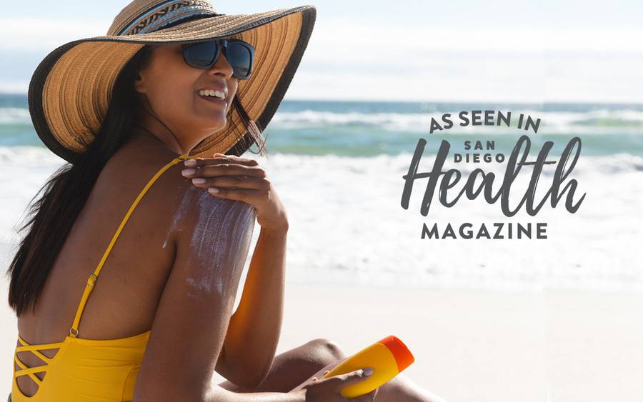 A young lady with tan skin applies sunscreen on her arm at the ocean - SD Health Magazine