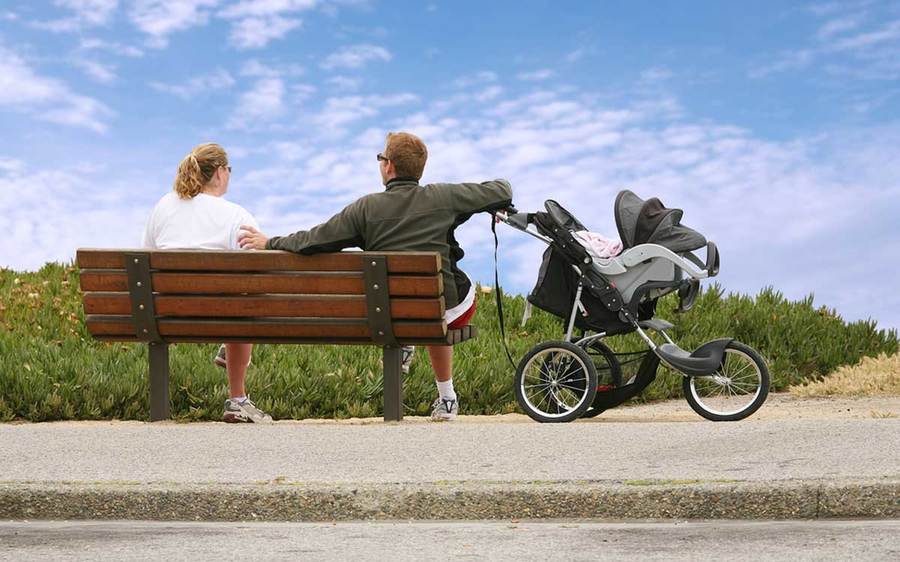 Two parents relax in the sunshine on a park bench while their baby stays safe in a covered stroller.