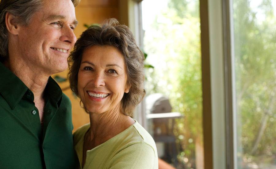 A couple smiles while standing together by a window in a sunlit room.