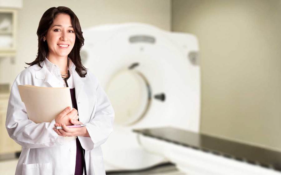 A professional interventional radiologist holding file folders stands before CT equipment.