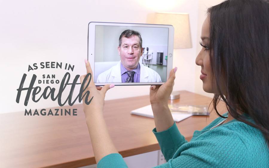 A young woman holds a tablet, watching and listening to her doctor on screen.