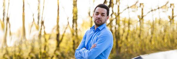 Man in blue shirt with goatee crossing his arms in front of backlit field.
