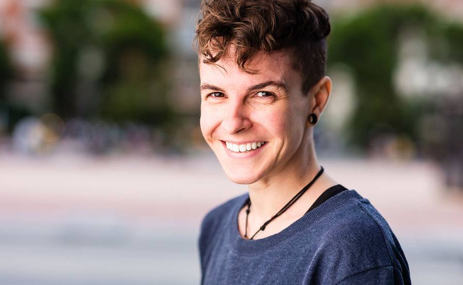 A young Caucasian person smiles at the camera near a busy park, comfortable in their gender identity.