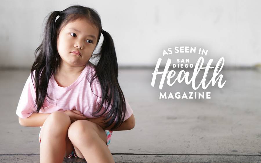 A young girl with pigtails sits on the ground with a look on her face that she doesn't feel well as she holds her stomach. SD Health Magazine