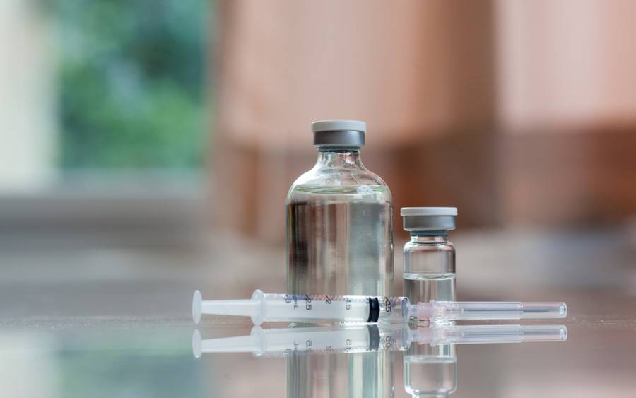 Two glass vials hold a clear vaccine fluid, next to a syringe.