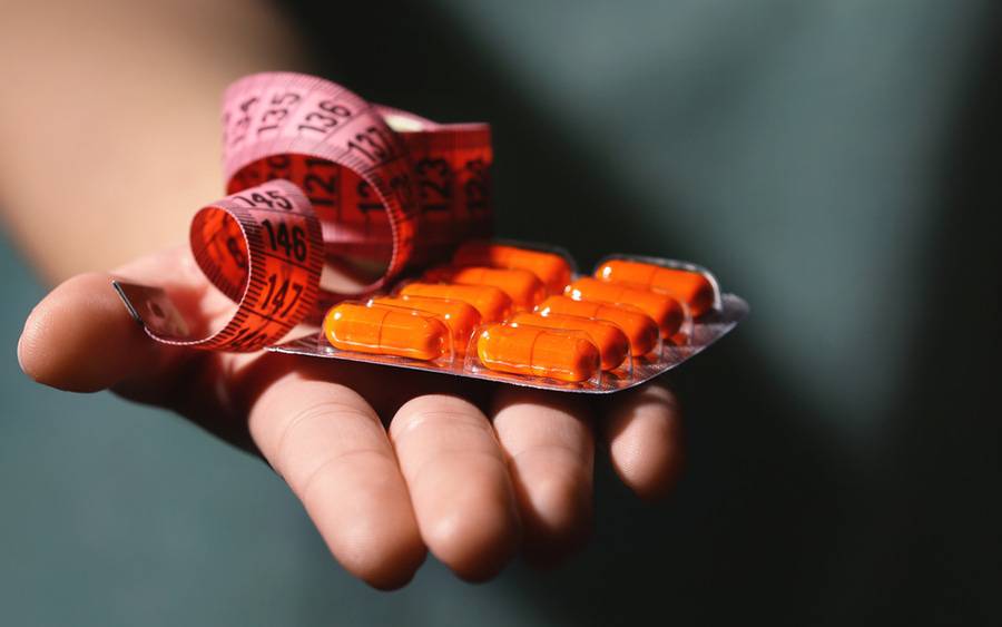 A closeup of a person's hand is showing off a new orange-colored weight loss pill and a tape measure.