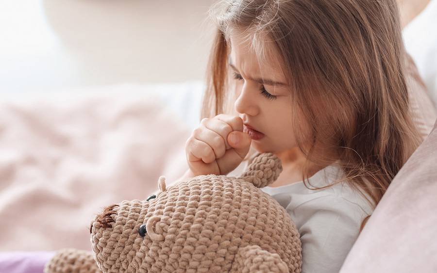 A little girl coughs in bed while  holding her play bear.