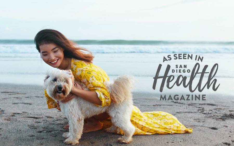 A woman affectionately pets her dog on the beach, illustrating the numerous physical and mental health benefits that come with pet ownership.