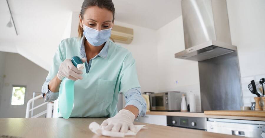 Woman with mask to protect against COVID-19 disinfects her kitchen table.