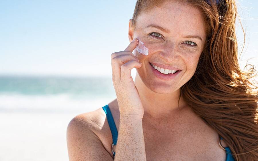 A freckled young woman smiles as she applies sunscreen on the beach.