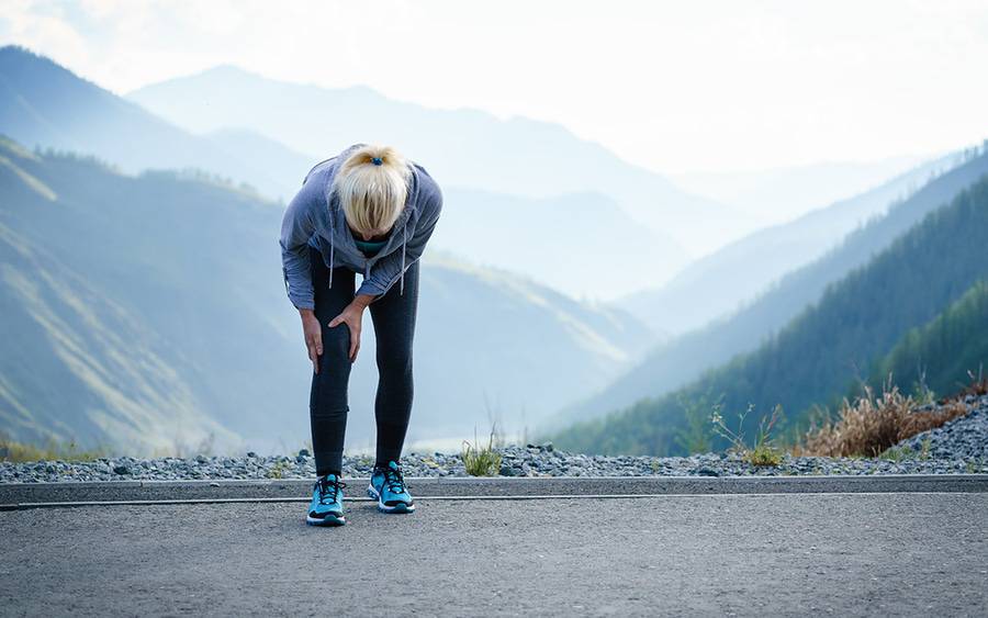 A woman bends over and grabs her knee in pain after running outside.