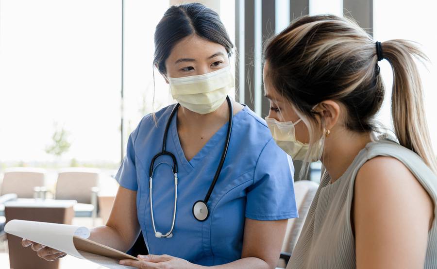 A medical professional wearing scrubs and surgical mask explains information from a clipboard to a patient.