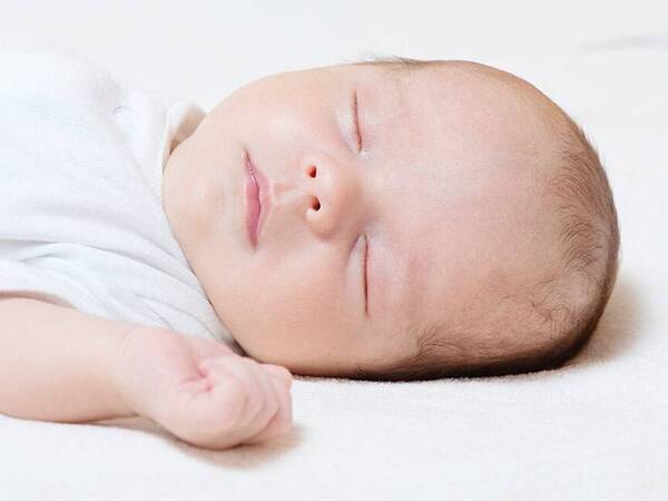 A newborn sleeps on its back, dressed in a white sleeping gown.