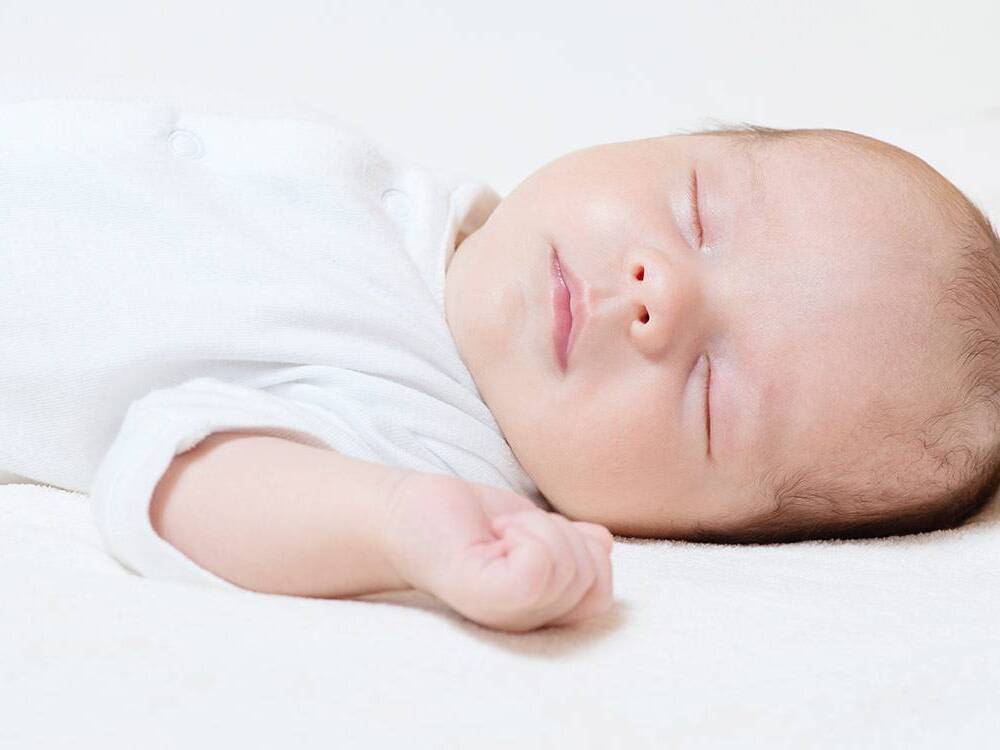 A newborn sleeps on its back, dressed in a white sleeping gown.