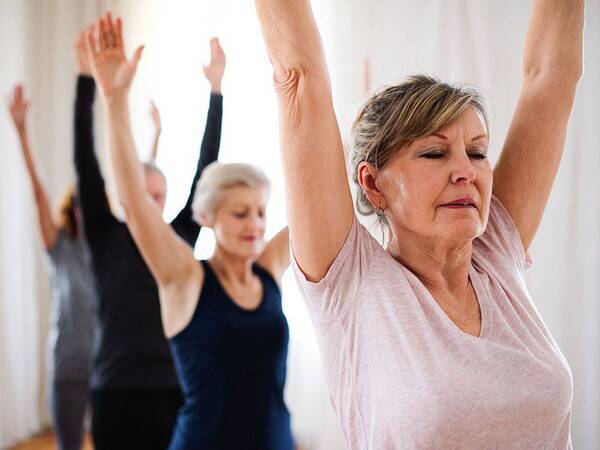 A group of people in an exercise class stand in a row with their arms stretched above their heads.