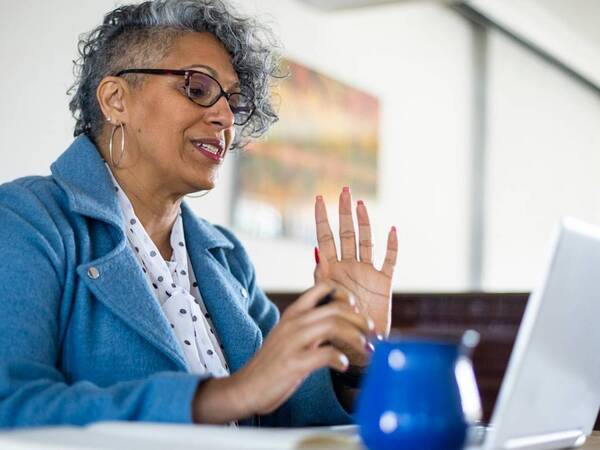 A woman with gray hair holds up her hand while watching a video on her laptop.