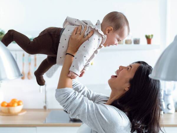 A woman in a kitchen gleefully raises an infant above her in the air.