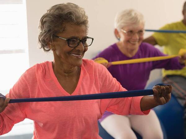A diverse group of older men and women smile while they participate in an exercise class using bands to improve their strength.
