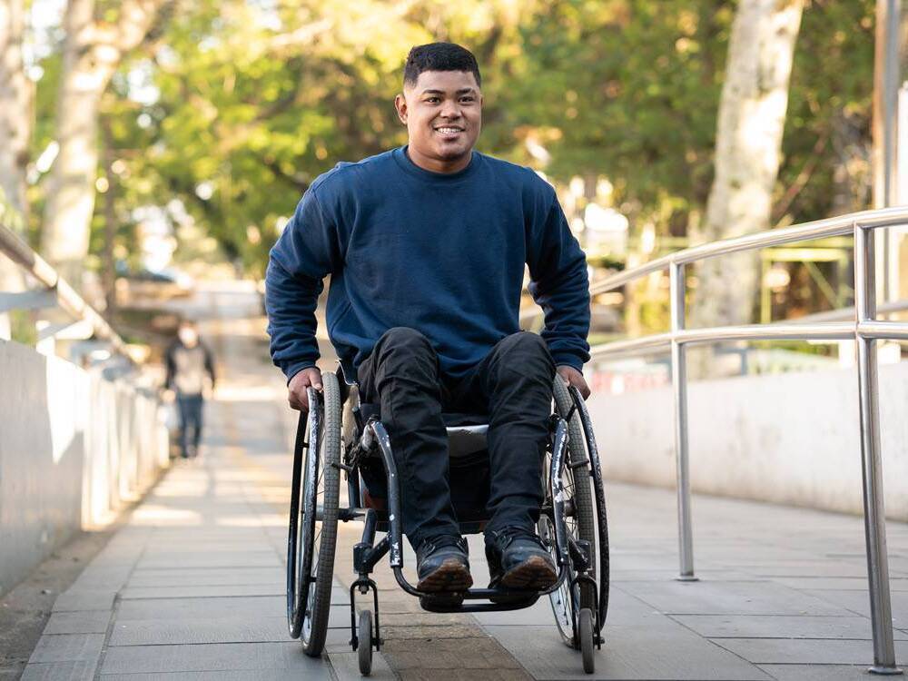 A young man in a wheelchair smiles as pushes himself up an outdoor ramp.