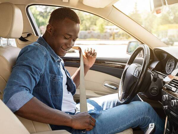 A young man smiles as he sits in the driver's seat of a car while he buckles his seat belt.
