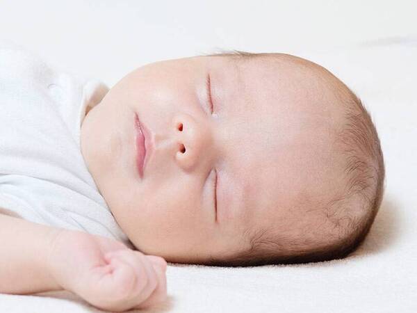 A newborn sleeps on its back, dressed in a white sleeping gown.