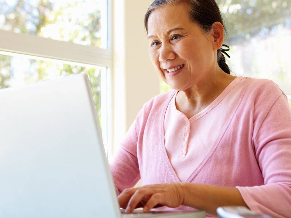A middle aged Asian woman wearing a pink shirt smiles as she sits in front of the laptop while participating in a virtual support group for advanced staged cancer.