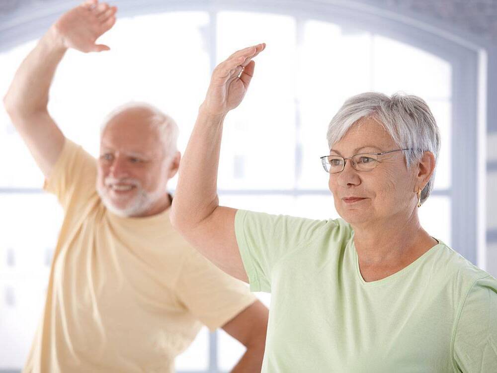A senior man and woman stretch their arm up in an exercise class designed to improve circulation and mobility.