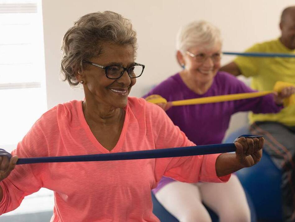 A diverse group of older men and women smile while they participate in an exercise class using bands to improve their strength.