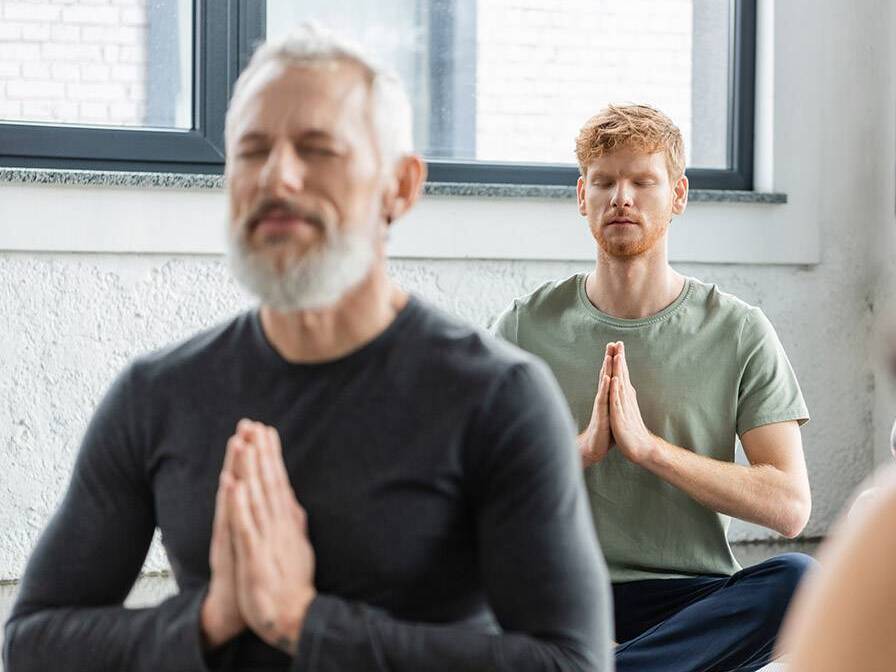 Men and women of various ages sit in a yoga pose with their eyes closed.