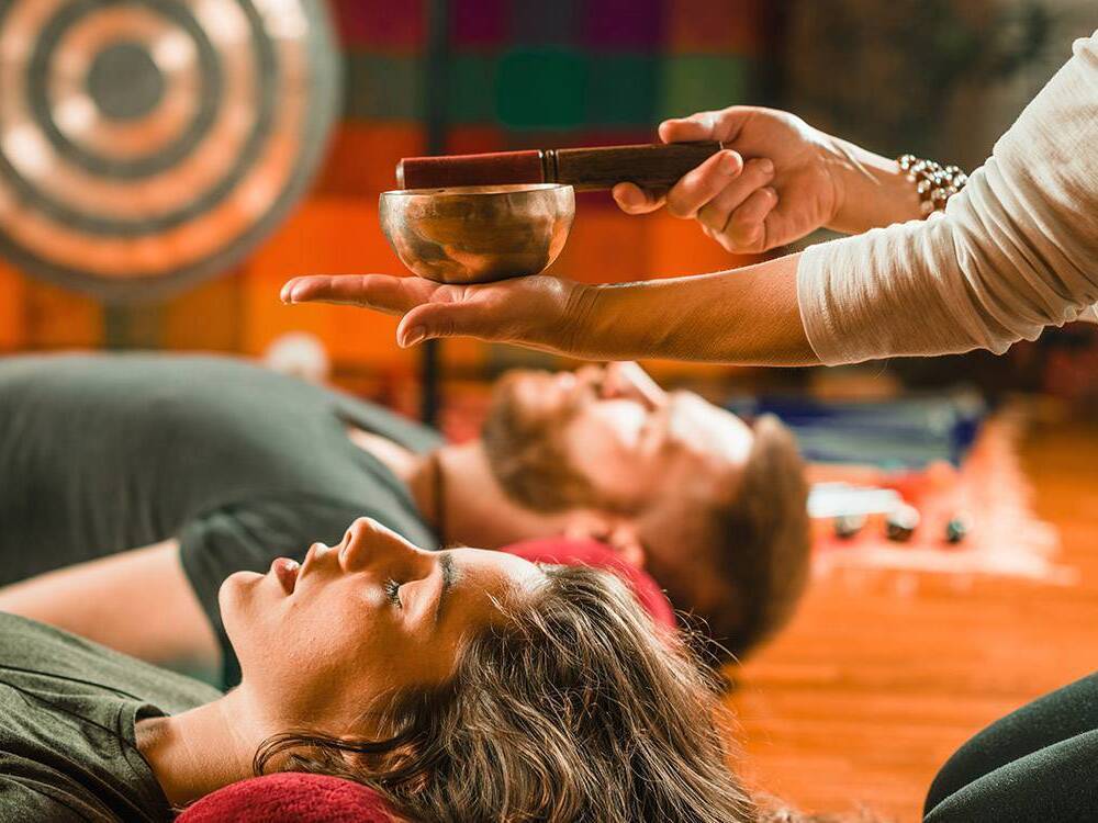 A yoga instructor sounds an instrument as participants relax comfortably with their eyes closed.