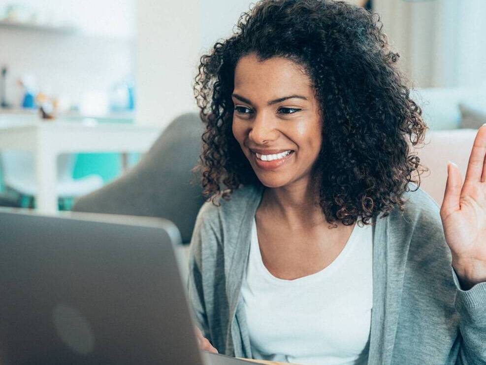 A middle-aged black woman smiles and raises her hand while participating in virtual caregiver support group on a laptop computer from her home.