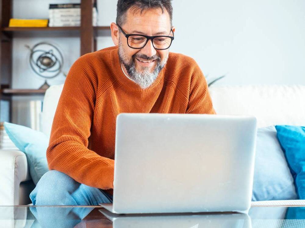 A man with gray hair smiles as he looks at a laptop while participating in a virtual support group for cancer survivors.