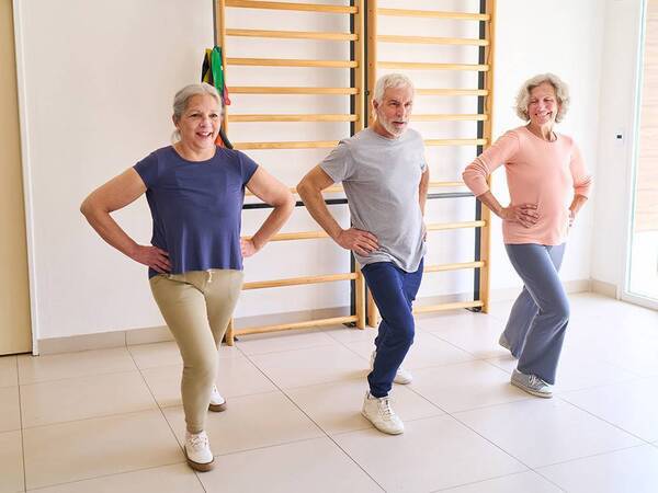Three senior men and women practice a standing stretch with one leg forward and slightly bent.