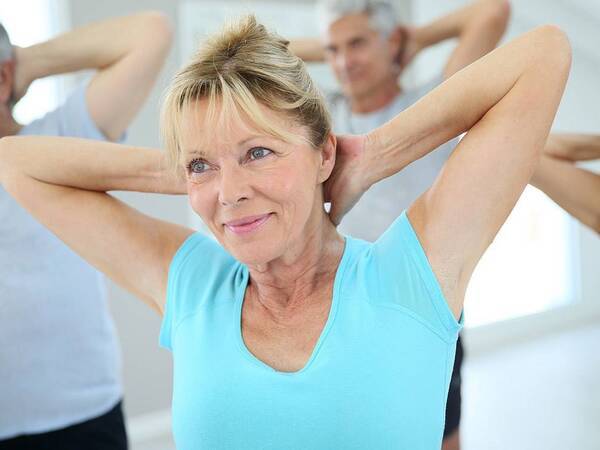 A senior woman with a blue shirt stretches with arms bent behind her head in an exercise class surrounded by other men and women.