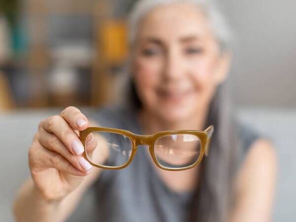 A woman with gray hair is blurred behind her eyeglasses that she's holding in front of her. 