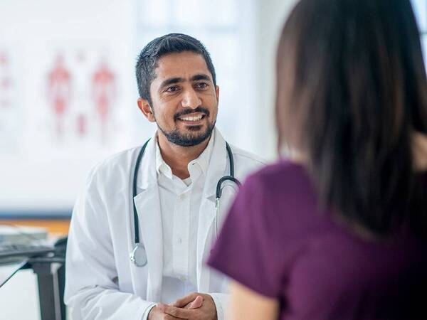 A physician speaking with a patient in an office with a computer and charts of the human body.