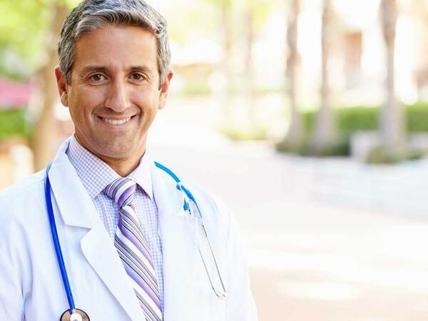 A physician stands in an outdoor setting with a white lab coat and blue stethoscope. 