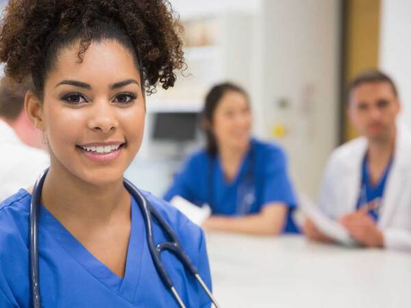 A medical professional wearing scrubs and a stethoscope takes a break from a conference table seated with other doctors and nurses.