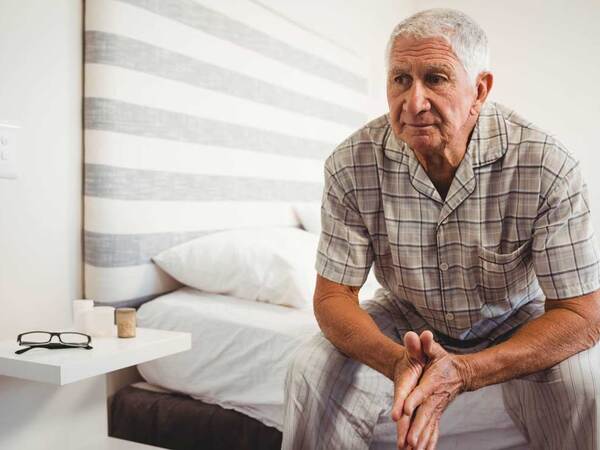 A thoughtful elderly man sits on his bed near a nightstand holding his glasses and candles. 