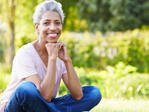 A smiling middle-aged woman wearing denim jeans sits cross-legged in a grassy park setting 