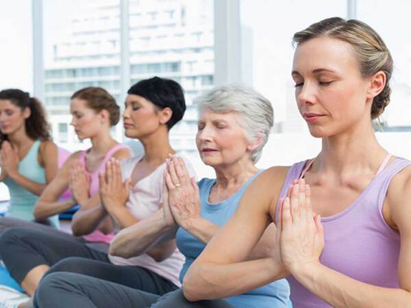 Five women of various ages seated in a yogic lotus pose with their eyes closed and palms pressed together, and  city skyline in the background.