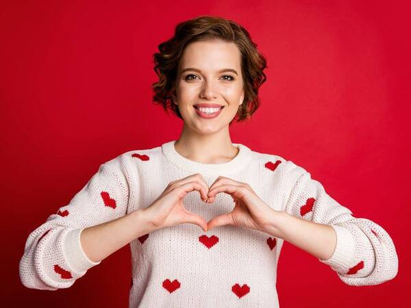 A woman smiles wearing a sweater with hearts. She makes a heart symbol with her hands in front of her heart.