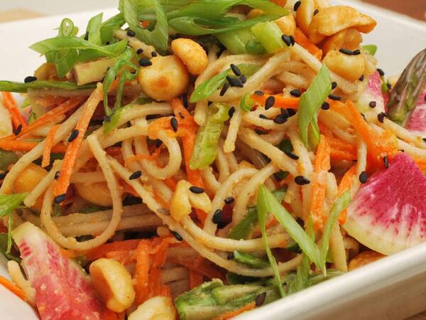 A dish of soba noodles with shredded carrots, asparagus tips, green onions, black sesame seeds and peanut sauce.